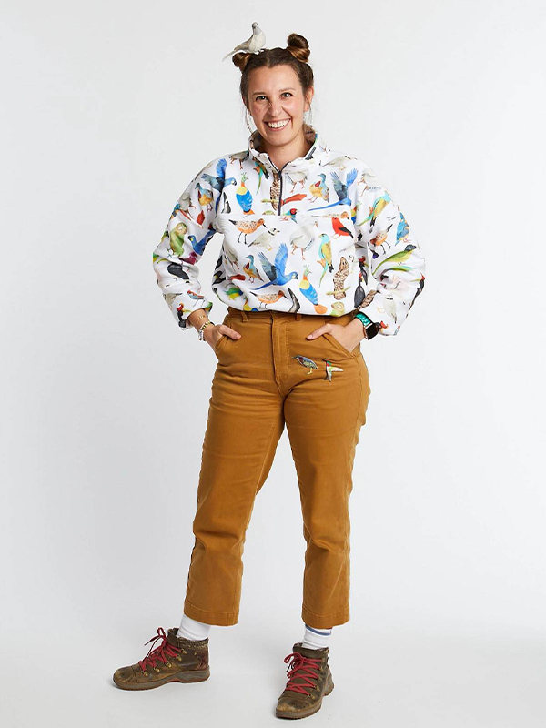 A woman wearing a pullover featuring a number of endangered birds and dark yellow pants stands in front of a white background. She is smiling at the camera and a small fake white dove is perched in her hair.