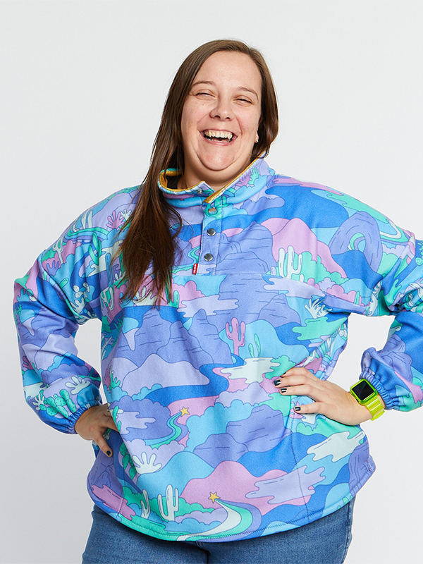 A woman wears a pullover featuring a turquoise, light blue and purple Arizona landscape design. She is smiling at the camera and standing in front of a white background.