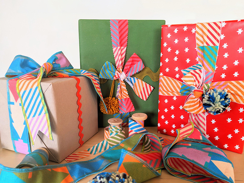 Holiday presents are wrapped in colorful fabric ribbon in jewel tone colors, in both a Christmas tree repeating design and small squares of coordinating colors in varying pairings.