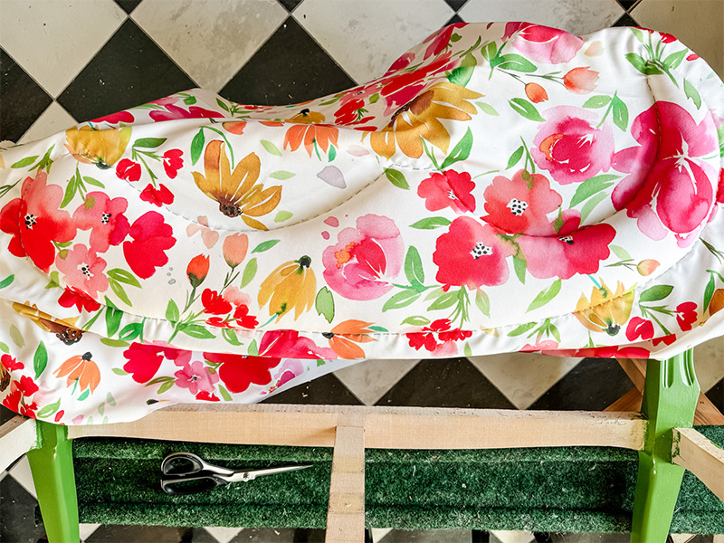 A floral fabric is stapled to the top of a settee. The excess fabric lays off the chair.