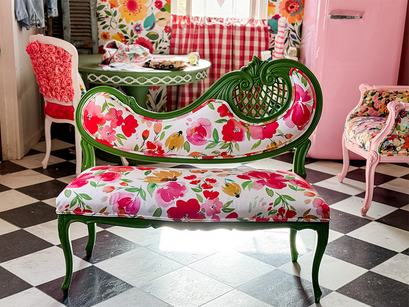 A green settee chair with floral fabric sits in the middle of a dining room with other upholstered chairs, a checked floor and pink fridge.