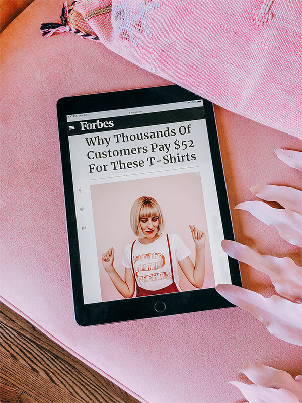 An iPad lays on a pink surface next to a pink pillow. On the iPad screen is the beginning of a Forbes article about Dani, with a photo of her wearing a shirt that says “Do the Damn Dream.” The headline of the article is “Why Thousands of Customers Pay $52 for These T-shirts.”