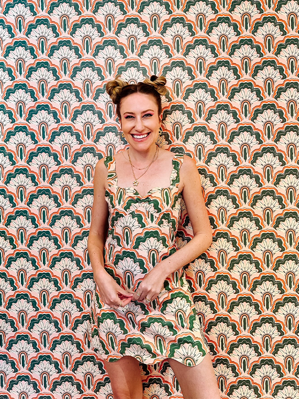 Dani stands in front of a wall featuring her wallpaper that has a scallop design in peach, green and white. She is wearing a dress sewn from fabric with the same design and is smiling.
