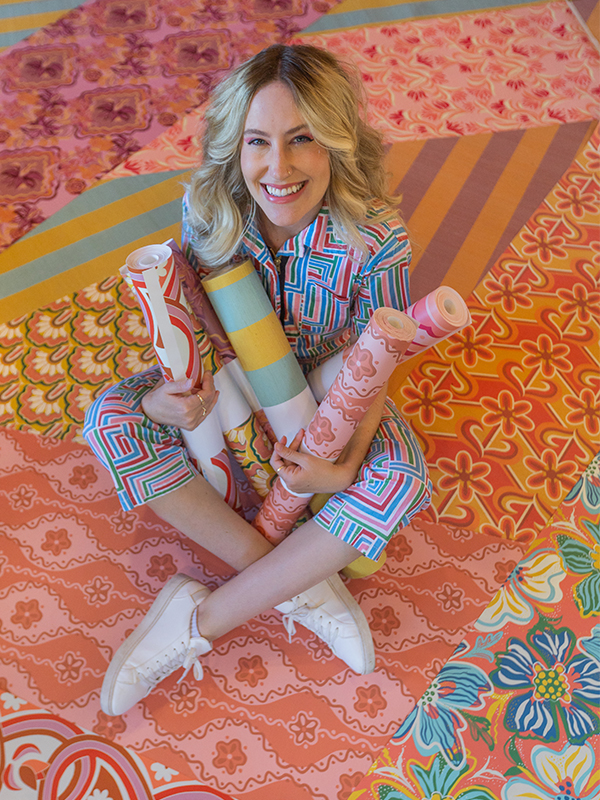 Dani sits cross legged on several different of her wallpaper designs laid out on the floor. She is also holding rolls of several of her wallpaper designs in her hands. She is wearing a bright geometric jumpsuit and looking up at the camera and smiling.