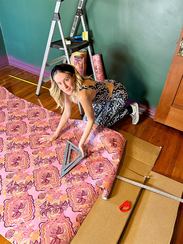 Dani kneels on the floor next to wallpaper that has been rolled on to the floor. She is holding a triangle ruler in her left hand and looking up at the camera. The wallpaper features a design with mauve flowers inside large squiggly diamonds on a purple background.