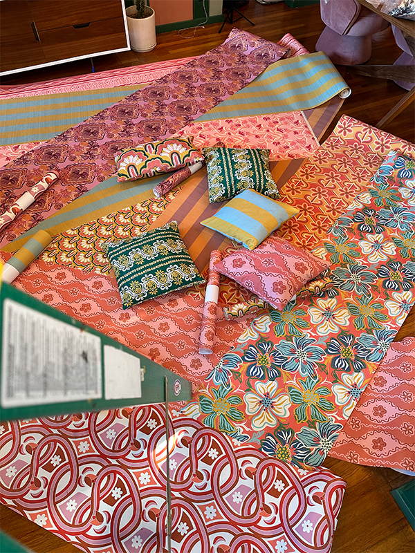 Several wallpaper rolls have been unrolled and laid out on the floor. Pillows and unrolled wallpaper rolls are also lying on the rolled-out wallpaper rolls. The designs on both the wallpaper and pillows are bright and cheery, most with pink tones. The photograph has been taken from the top of a ladder, the legs of which are visible on the left-hand side of the photo.