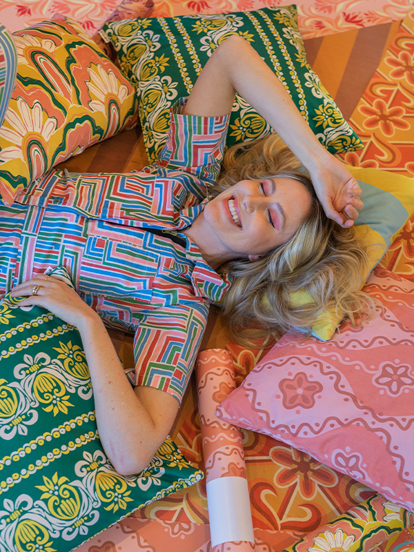 Dani is laying on top of several unrolled rolls of wallpaper and throw pillows, all featuring her designs, laid out on the floor. She is wearing a bright geometric jumpsuit and smiling and has her eyes closed.