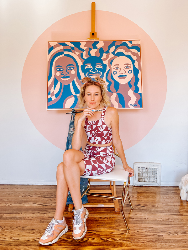 Dani sits on a small table in front of a painting of hers featuring three people in blue, mauve and pink. The painting is hung on a wall in the middle of a large painted pink circle. Dani is holding a paintbrush parallel to her chin and looking at the camera.