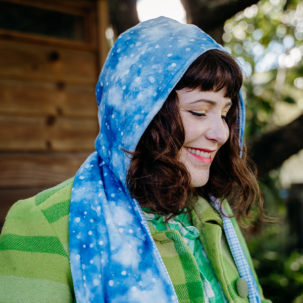 A close-up side view a hooded scarf. The fabric is blue with small white circles in different sizes, like snowflakes. The model is wearing a green and white plaid coat, smiling and looking down.