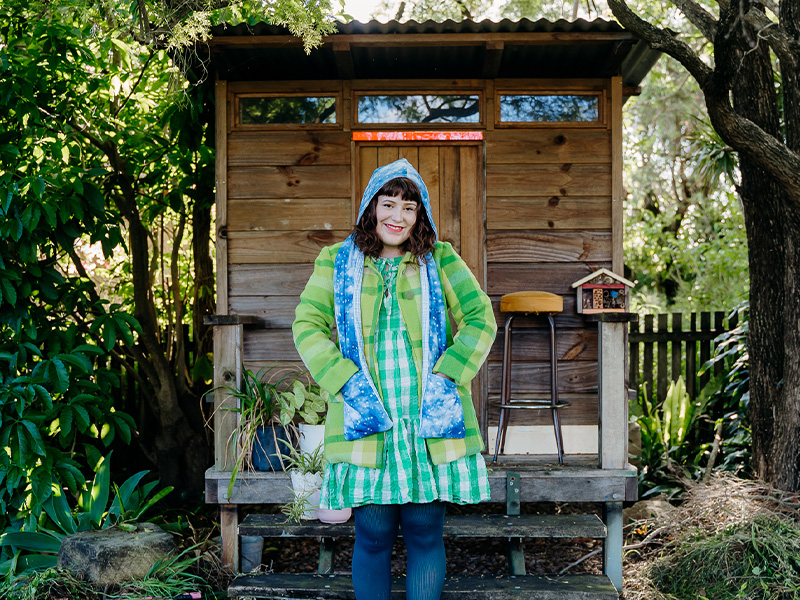 Woman in a green jacket and dress, blue tights and green boots poses in front of a small house surrounded by foliage is wearing a blue hooded scarf.