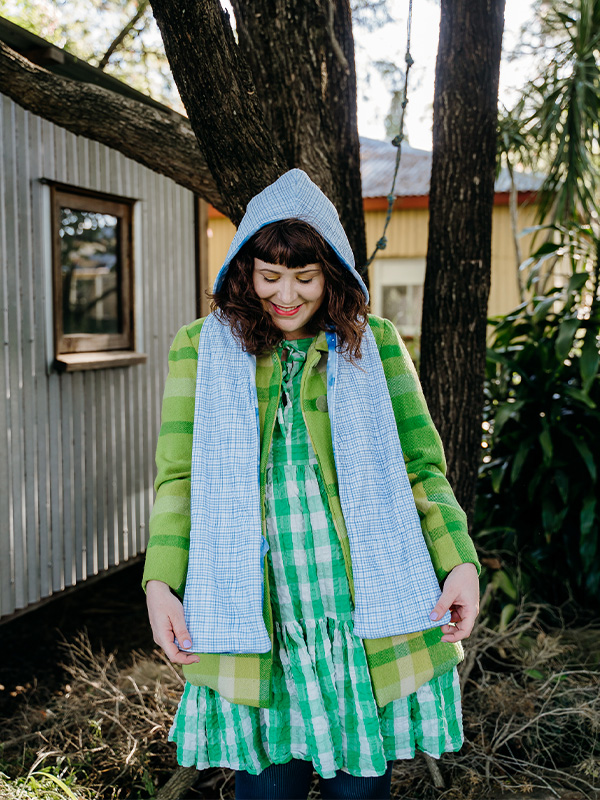Woman wearing green jacket and dress admires her blue and white hooded scarf by holding the ends of the scarf looking down at them. A house and trees are in the background.