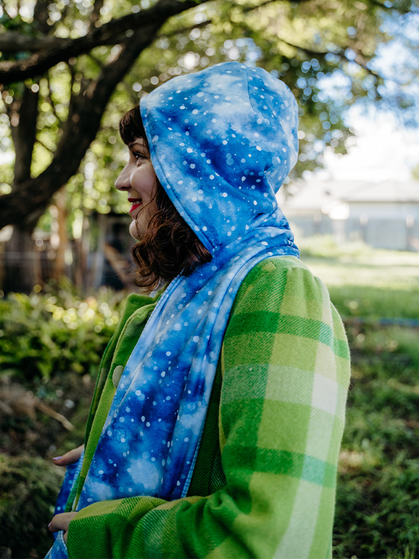 A profile of a woman wearing a green coat a blue and white hooded scarf. Trees and grass are in the background.