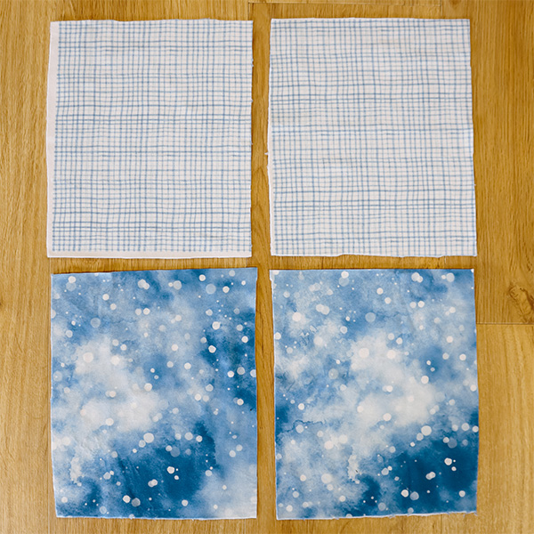 Four fabric squares lay on a wooden surface. Two are a blue grid design, two are a blue galacy like design.