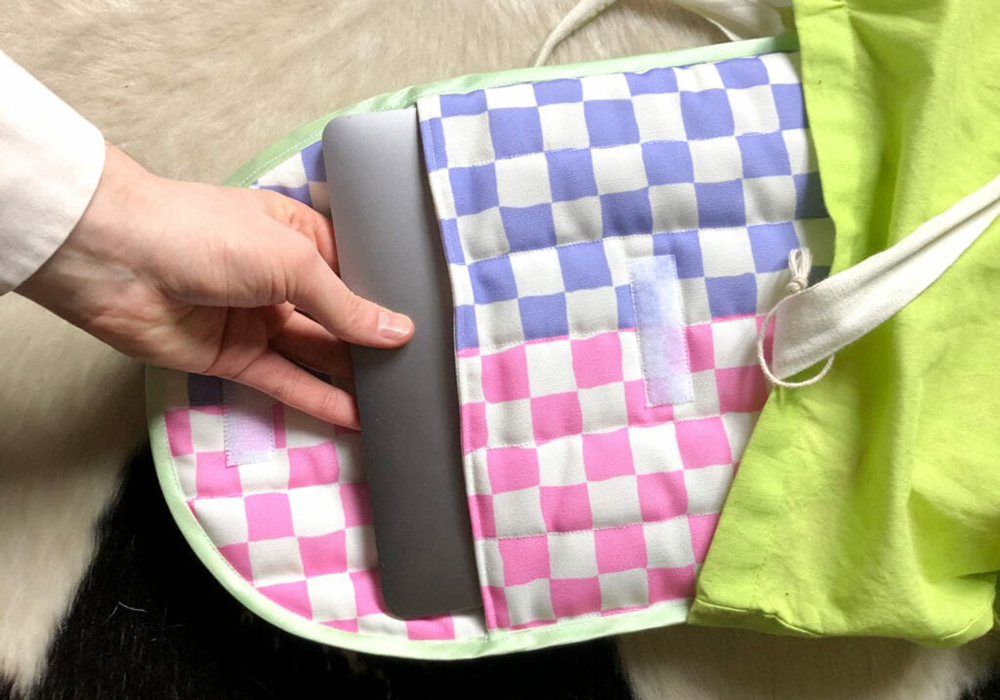 A hand is pulling out a silver laptop from a quilted laptop bag. The bag's fabric is pink and white checkerboard and purple and white checkerboard.