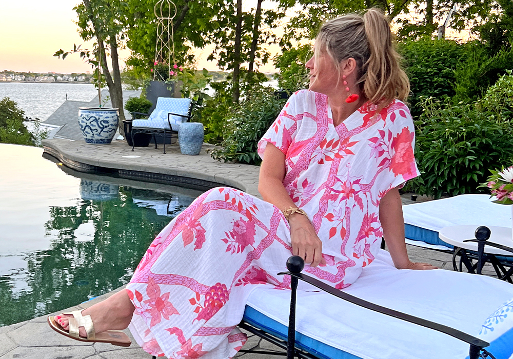 Danika sits by a pool in a V-neck caftan with hot pink flowers on a white background.