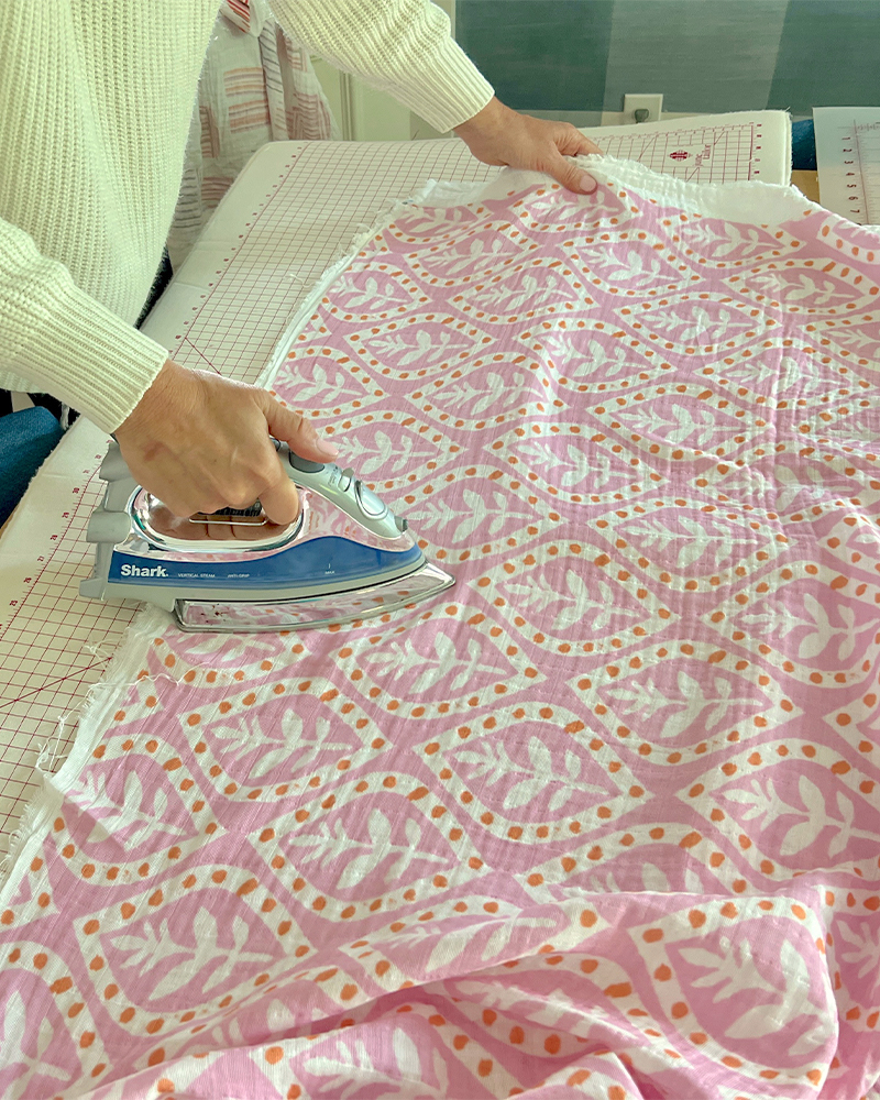 Pressing fabric for the caftan with an iron. The fabric design is repeating white flowers on a pink background. Each flower is encircled by a white oval with orange dots.