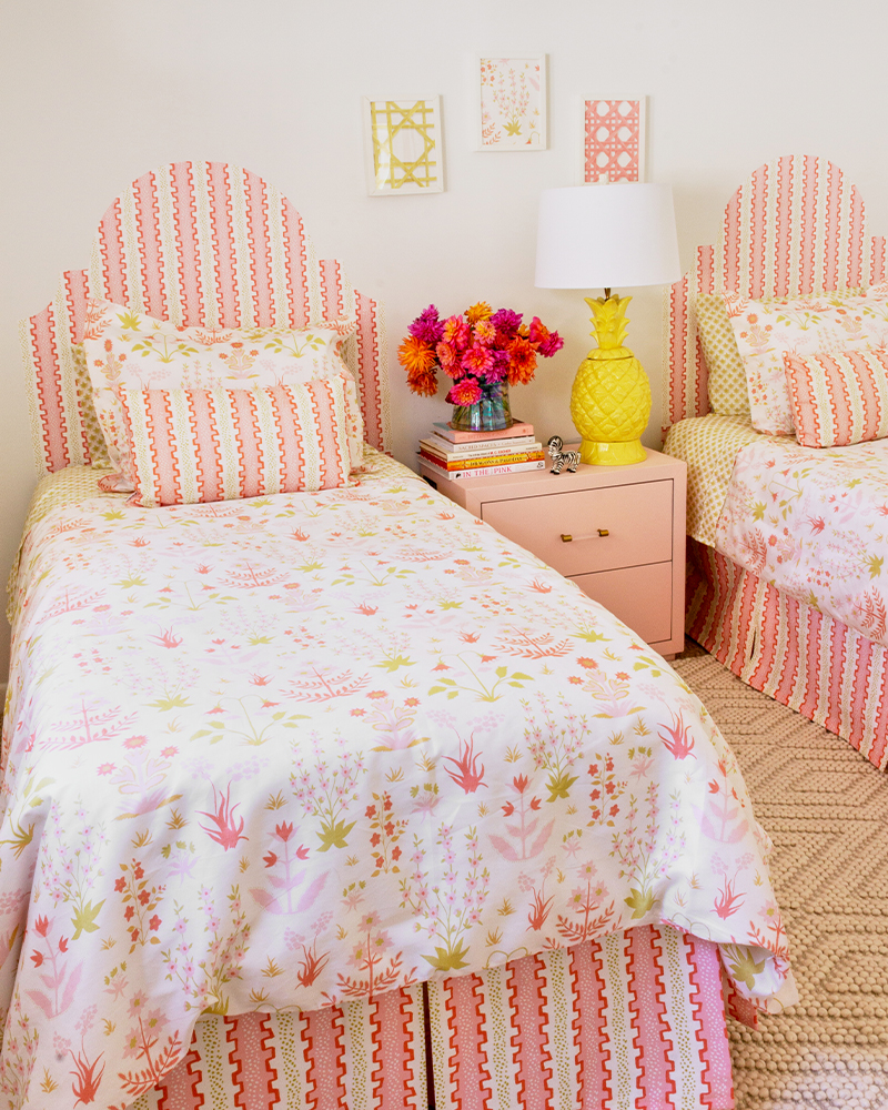 A look from the left of the finished room. The designs on the art, bedding and headboards are light pink and green florals. A pink bedside table with a bouquet of bright flowers to the left and a yellow lamp to the right is between the two beds.