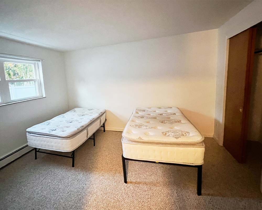 The dorm room before being revamped by Danika. Two twin beds sit side by side in a room with tan carpet and beige walls. A long horizontal window is to the left, a closet with a dark brown wooden door is to the right.