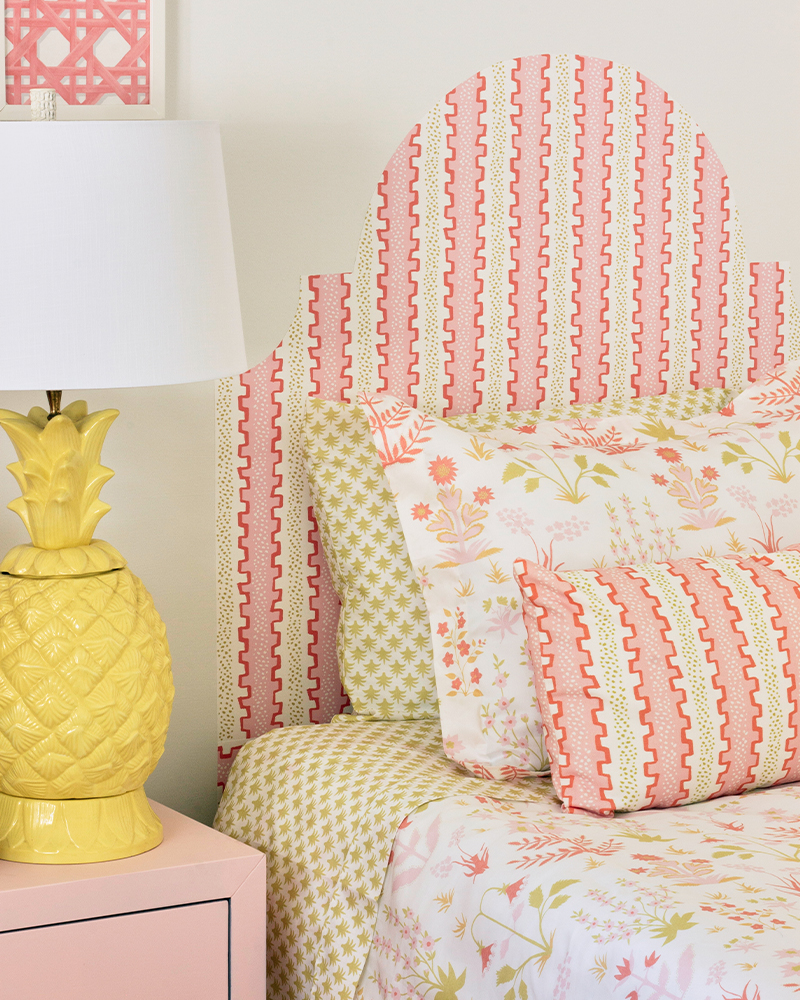A close up of the finished headboard and the top of the made-up bed, along with a piece of artwork to the top left. A yellow lamp has been placed on a pink bedside table to the left of the bed. The designs on the art, bedding and headboard are light pink and green florals.