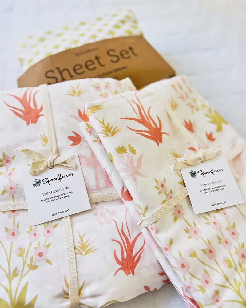 A close up of the duvet covers and one of the sheet sets. The sheet set fabric pattern is white with small green floral sprigs. The duvet cover fabric pattern features pink flowers and green leaves on a cream background.