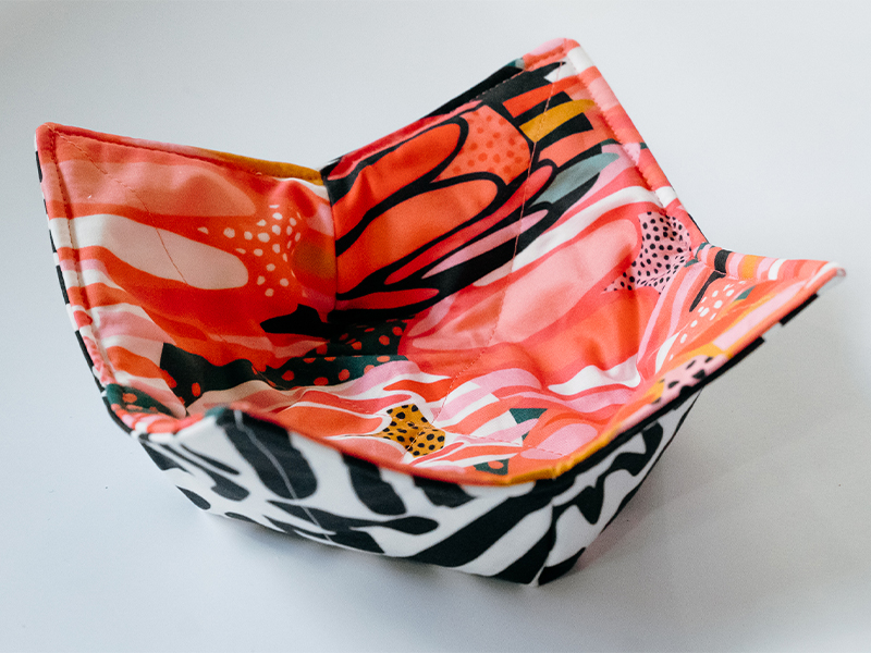 The finished bowl cozy as seen from the right side. It has a concave shape to fit a bowl of soup and four pointed edges. The fabric on the top side features bright flowers in inks, reds, yellows and white. The fabric on the bottom side features black geometric lines on a white background.