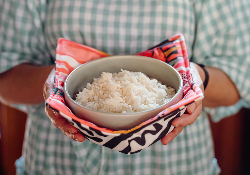 A close up of a person wearing a blue-and-white gingham dress holds a bowl of rice that itself is held in a fabric bowl cozy. The fabric on the top side of the cozy features bright flowers in inks, reds, yellows and white. The fabric on the bottom side features black geometric lines on a white background.