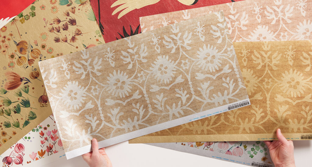Two hands are held apart, each holding a different wallpaper 2’ x 1’ swatch featuring a design with cream flowers and stems on a light brown background. The left hand holds a Silver Metallic Wallpaper swatch where the cream elements show up on a silver background. The right hand holds a Gold Metallic Wallpaper swatch where the cream elements show up on a gold background. Lying on the white surface below both hands are other Metallic Wallpaper samples, some with watercolor jewel-tone flowers and some with red cranes.